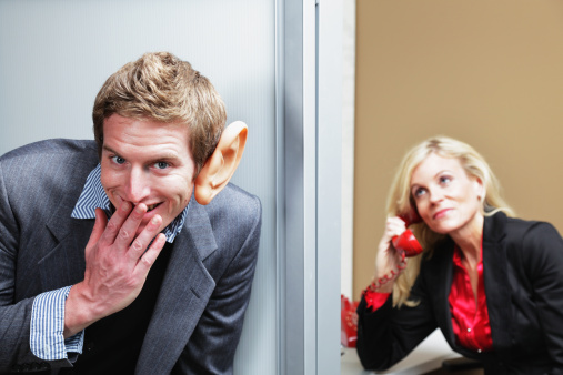 A young man with an exaggeratedly large ear is smirking as he eavesdrops outside a woman's office.