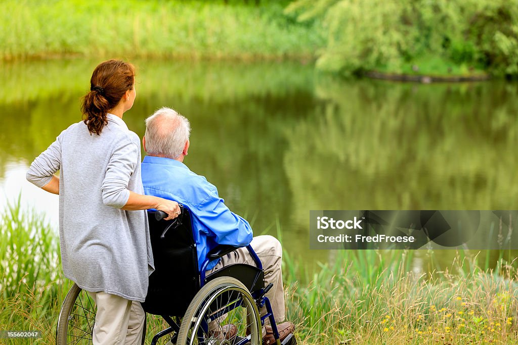 Sênior homem sentado em uma cadeira de rodas com cuidadora - Foto de stock de 70 anos royalty-free