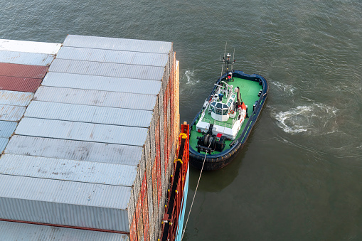 High angle view of the stern of a large container ship loaded with containers while being towed by a tugboat, rope attached to ship
