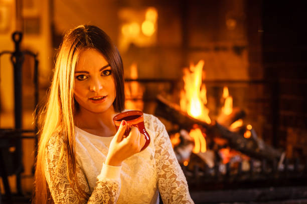 Woman drinking cup of coffee relaxing at fireplace. Young girl with hot beverage heating warming up. Winter at home.