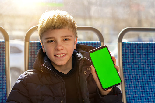 Happy teen boy sits looks at camera and shows smartphone with green screen for new application. Excited kid rides on tram and presents phone with chromakey to camera.