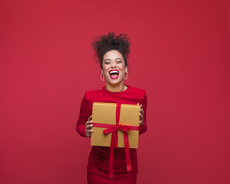 Young woman wearing red elegant dress, holding Christmas present and laughing at camera. Studio shot, red background.