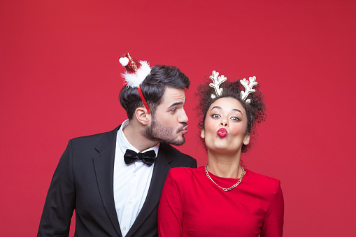 Young woman and man wearing elegant clothes and Christmas headbands blowing kisses. Studio shot, red background.