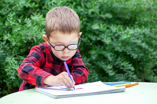 A little boy draws in the garden (outdoors). He wears glasses and an eye patch (occluder) to prevent amblyopia and strabismus (squint, lazy eye). The school students vision diseases problem.
