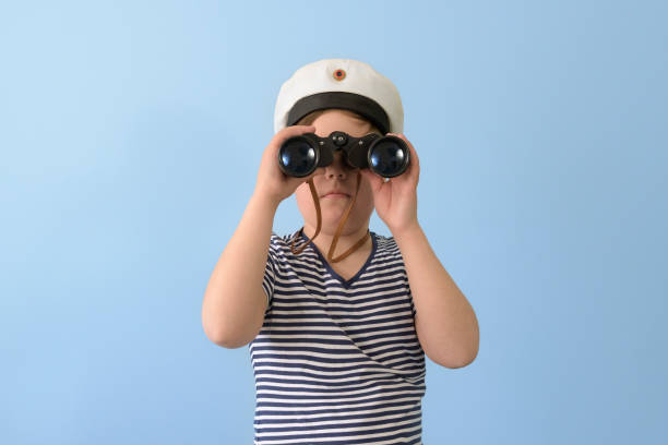 Caucasian child in blue T-shirt looking at camera with binoculars hanging from neck with surprised expression, on white background.