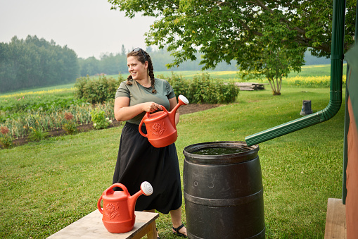 Smiling young woman filling watering cans with rainwater from a catchment system to water her vegetable garden on a farm