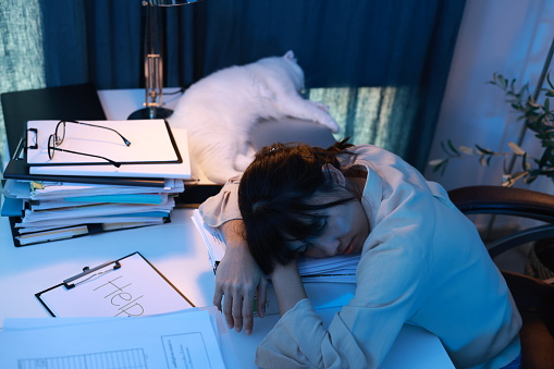 The woman's office was very tired and fell asleep at the workplace, her head lying on the table. A lot of paperwork. Tired employee, a stack of documents and papers. Sleepy tired alone girl dozing off at the table while working hard at late night.