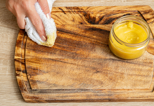 Woman hands apply homemade beeswax wood treatment polish to restore natural wood cutting board. Beeswax, olive oil and essential oil, soft cloth and mixture in glass jar. Polishing wood.