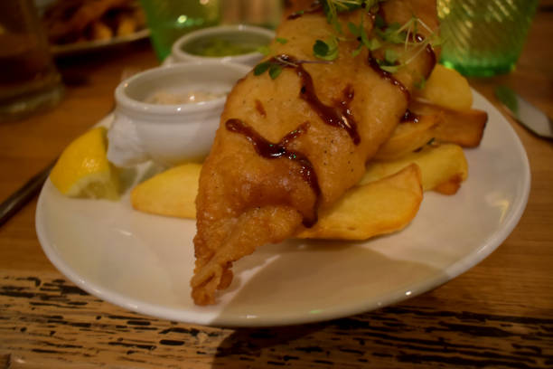 A side viewed of white plate of Fish and chips. A subtle Asian take on a classic with lightly-battered haddock, salt-pepper chips, chilli & lime crusted peas, served with garlic and oriander tartare sauce. una vista lateral de plato blanco de pescado y patatas fritas. una sutil versión asiática de un clásico con eglefino ligeramente rebozado, chips de sal y pimienta, guisantes con costra de chile y lima, servidos con ajo y salsa tartar de oriandro.