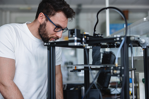 A young male designer wearing glasses is looking with attention a 3D printer working. The orinter is modelling a figure.