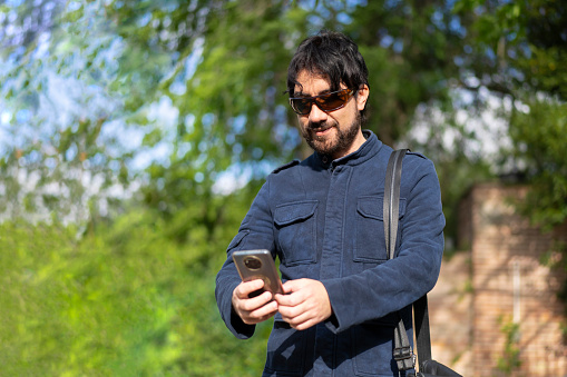 Happy young handsome Hispanic hipster man using phone at the park