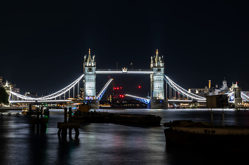 Illuminated Tower Bridge lifting a drawbridge for a seagoing vessel crossing the River Thames at night in London.