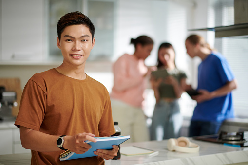 Portrait of cheerful college student with textbook, his friends talking in background