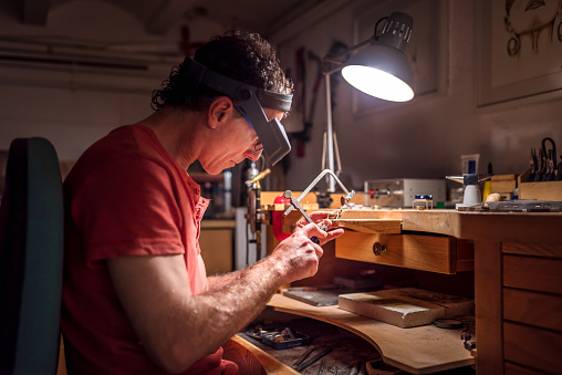 side view portrait of a male Caucasian jeweller sawing metal in his workshop. Jewel, craft and luxury concept.