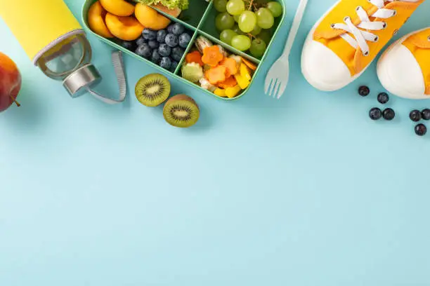 Photo of Nourish your brain: Top-down view of sneakers, a lunch box packed with sandwiches, fruits, vegetables, and a water bottle on a pastel blue isolated backdrop, ready for text or promotional content