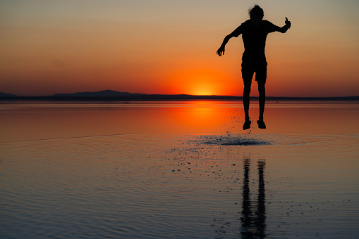 Young male bouncing on the water surface in Salt Lake. The sun is setting on the water horizon. The reflection of the young man jumping into the air falls into the water. Shot with a full-frame camera in daylight.