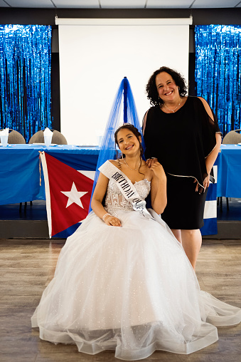 Quinceanera for hispanic teenager posing with mother. She is dressed with beautiful party gown, honouring her cubans origins and traditions. She has long brown hair and braces. Vertical full length indoors shot with copy space. This was taken in Quebec, Canada.