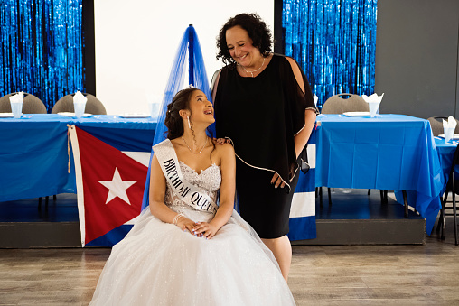 Quinceanera for hispanic teenager posing with mother. She is dressed with beautiful party gown, honouring her cubans origins and traditions. She has long brown hair and braces. Horizontal waist up indoors shot with copy space. This was taken in Quebec, Canada.