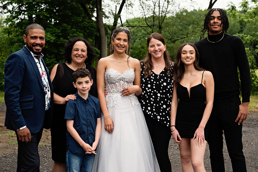 Quinceanera for hispanic teenager posing with family. She is dressed with beautiful party gown, honouring her cubans origins and traditions with her multiracial family. She has long brown hair and braces. Horizontal mid length outdoors shot with copy space. This was taken in Quebec, Canada.
