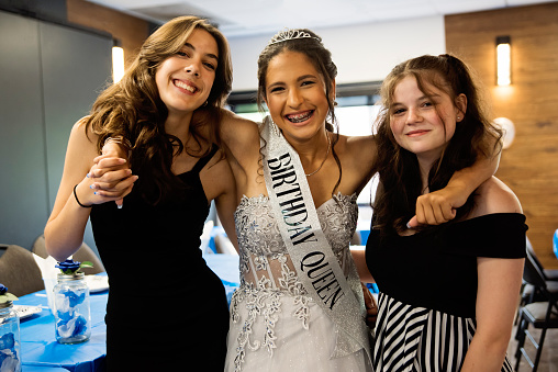 Quinceanera for hispanic teenager posing with best friends. She is dressed with beautiful party gown, honouring her cubans origins and traditions with her two best friends. She has long brown hair and braces. Horizontal waist up indoors shot. This was taken in Quebec, Canada.