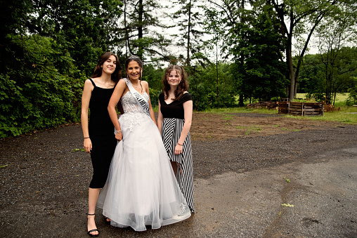 Quinceanera for hispanic teenager posing with best friends. She is dressed with beautiful party gown, honouring her cubans origins and traditions with her two best friends. She has long brown hair and braces. Horizontal full length outdoors shot with copy space. This was taken in Quebec, Canada.