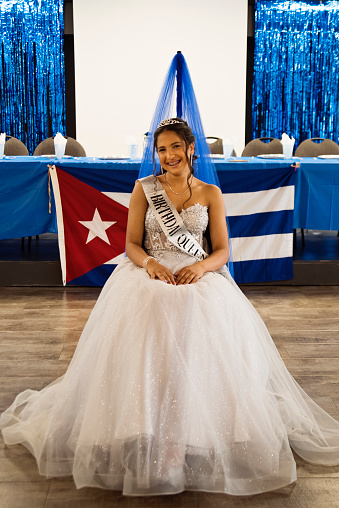 Portrait of beautiful hispanic teenager posing for Quinceanera. She is dressed with beautiful party gown, honouring her cubans origins and traditions. Cuban flag in the background. She has long brown hair and braces. Vertical full length indoors shot with copy space. This was taken in Quebec, Canada.