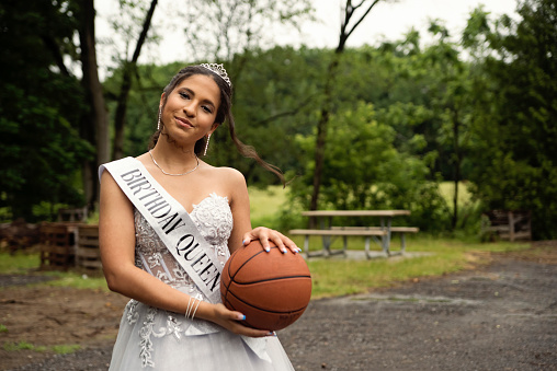 Portrait of beautiful hispanic teenager posing for Quinceanera. She is dressed with beautiful party gown, honouring her cubans origins and traditions. She has long brown hair and braces and is holding her beloved basketball. Horizontal waist up indoors shot with copy space. This was taken in Quebec, Canada.