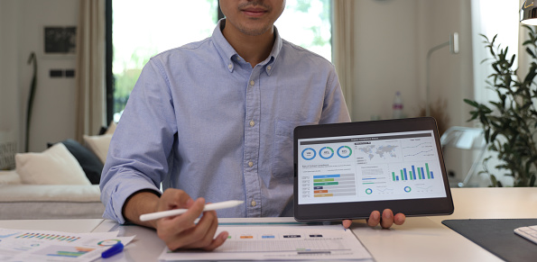 Young businessman sitting at office desk looking at camera and pointing at a tablet with financial information displayed in graphical form (column graph, stacked bar graph, and pie graph).