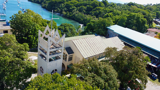 Church building and bell tower with a cross in a small Asian town. Aerial view of the catholic church on the seashore among dense green trees.