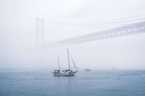 View of 25 de Abril Bridge famous tourist landmark of Lisbon connecting Lisboa and Almada in heavy fog mist wtih yacht boats passing under. Lisbon, Portugal