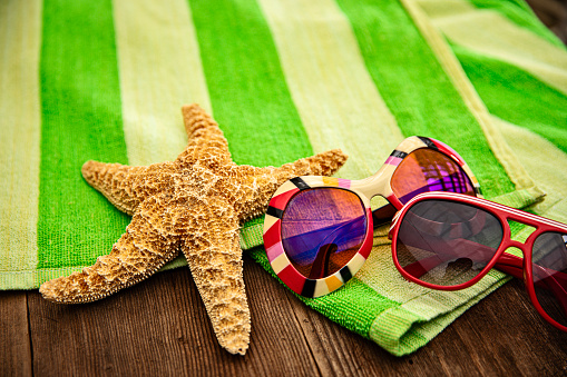 This is a close up photograph of a pair of sunglasses. One is for men the other is for women sitting on top of a green striped beach towel on a wood deck. There is space for a copy