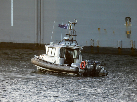 A motorboat of the Australian Federal Police patrols Sydney Harbour where USS Canberra (LCS 30), an Independence Class combat ship of the US Navy, is docked at Garden Island.  The USS Canberra is in port for a commissioning ceremony on 22 July 2023.  In the background is the hull of HMAS Canberra of the Royal Australian Navy, docked next to her.  This image was taken from Mrs Macquarie's Chair on a cold, sunny and windy morning on 19 July 2023.