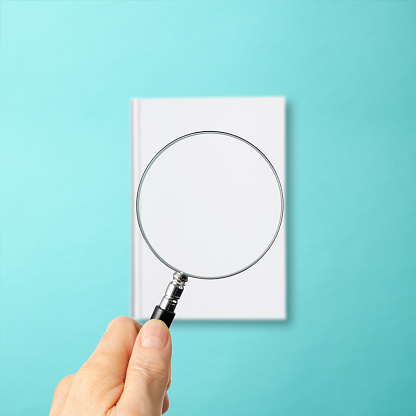 Overhead shot of examining a closed blank book with a magnifying glass on light blue background.
Focus on Foreground.