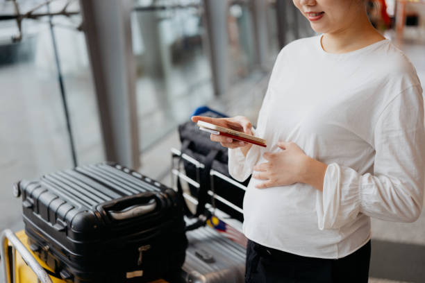 Pregnant Asian woman using smartphone in the airport Image of a pregnant Asian Chinese woman using smartphone in the airport. Pregnant woman online check in with smartphone for electronic boarding pass. asian pregnant woman airport stock pictures, royalty-free photos & images