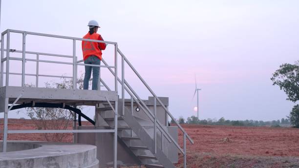Asian female environmental engineering wear safety uniform, working in station at wind turbine fields to generate electricity, checks the status of the turbines using a tablet. Renewable energy for sustainable development. Power generation from natural energy. las ingenieras ambientales asiáticas usan uniforme de seguridad, trabajan en estaciones en campos de turbinas eólicas para generar electricidad, verifican el estado de las turbinas usando una tableta. energía renovable para el desarrollo sostenible. gen