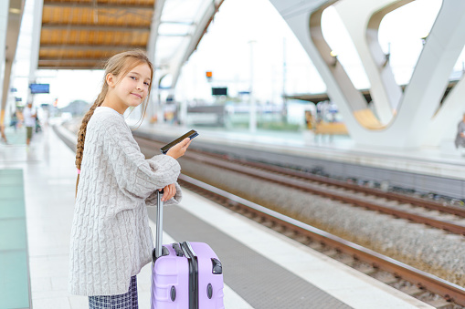 Little adorable girl with suitcase is waiting for ride at station while waiting for train.