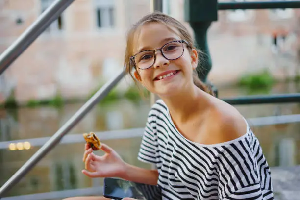 Portrait of little charming happy girl in glasses sits on bund of canal river in summer.