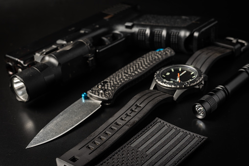 A flat lay with various black EDC (everyday carry) items on a black background. Including a pocket knife, hand gun, watch, flash light and US flag patch.