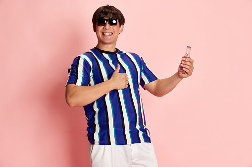 Portrait of young smiling, excitement man in sunglasses standing with bottle and showing gesture of good against pink background. Concept of human emotions, lifestyle, casual fashion, positivity, ad