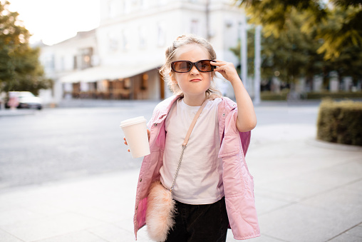 Cute kid girl 4-5 year old wear sun glasses and trendy casual clothes holding coffee cup having fun at city street over sunset outdoor. Portrait of cheerful and happy child. Summer season.