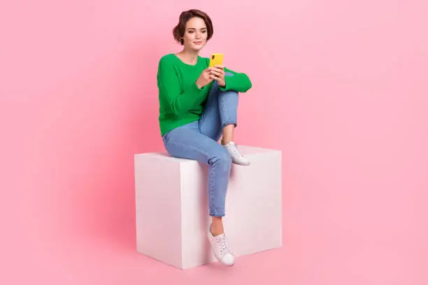 Photo of Full length photo of adorable girl wear stylish clothes sit cube use modern technology samsung iphone isolated on pink color background