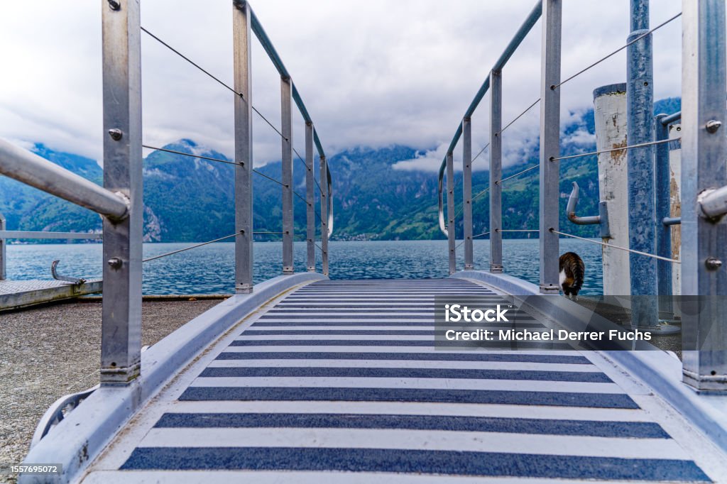 Diminishing perspective of gangway with Lake Lucerne in the background. Scenic view of Lake Lucern with gangway at pier of village Sisikon at on a cloudy spring day. Photo taken May 18th, 2023, Sisikon, Canton Uri, Switzerland. Aisle Stock Photo
