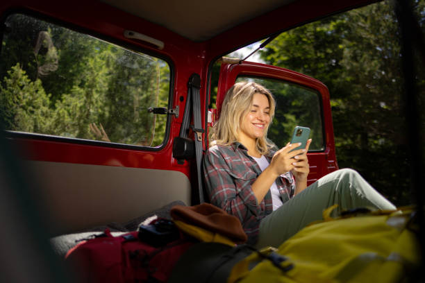 istock Happy female backpacker text messaging on cell phone while relaxing in her van. 1557721779