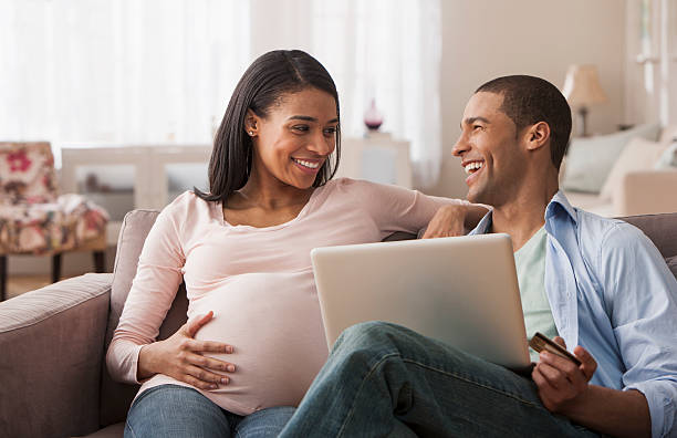 mujer embarazada joven sentado con esposo y computadora portátil - home finances couple computer african ethnicity fotografías e imágenes de stock