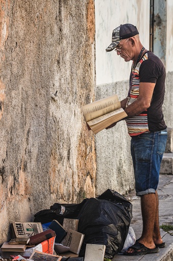 Matanzas, Cuba – May 08, 2023: A mature male holding an old book near a trash pile of books