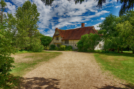 A large English Tudor Farm house with a gravel drive grass lawn and trees. The house has a red tiled roof and the scene is a summers day with blue sky and white clouds