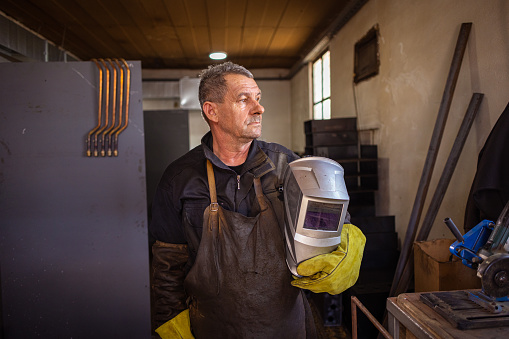Portrait of a mature welder, standing in his metal shop, holding welding helmet.