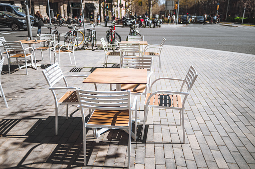 Barcelona; Ample sidewalk space adorned with empty, inviting white and brown tables and chairs, perfectly spaced for visitors to savor moments while bicycles rest nearby, a captivating spot to unwind
