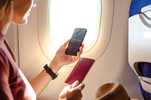 Young woman sitting on the aircraft seat near the window during the flight in the airplane. She is using smart and holding passport