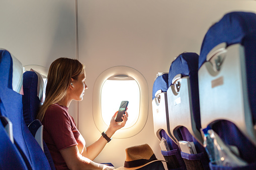Young woman sitting on the aircraft seat near the window during the flight in the airplane. She is using smart phone and watching online content